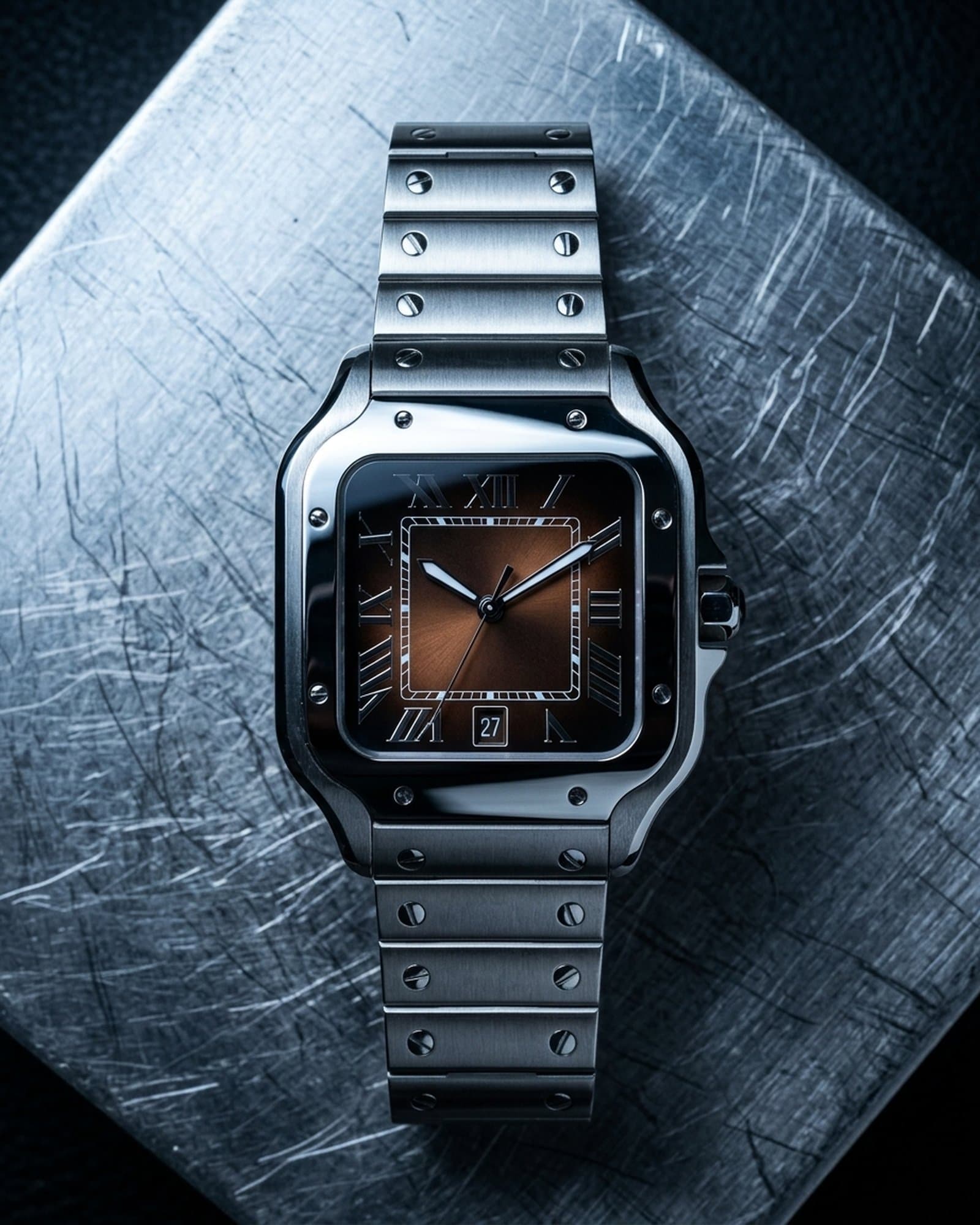 Square watch with a brown dial lying on scratched steel under a cold light