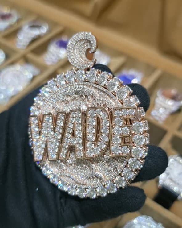 Round rose gold medallion with a name in raised letters over a pavé disc and a ring of large stones, held in a black glove